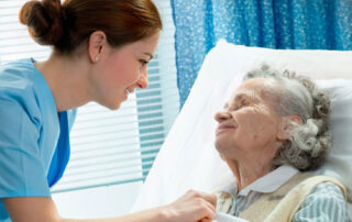 A female nurse in a blue uniform is fixing the sheet on an elderly woman who is lying in a hospital bed.