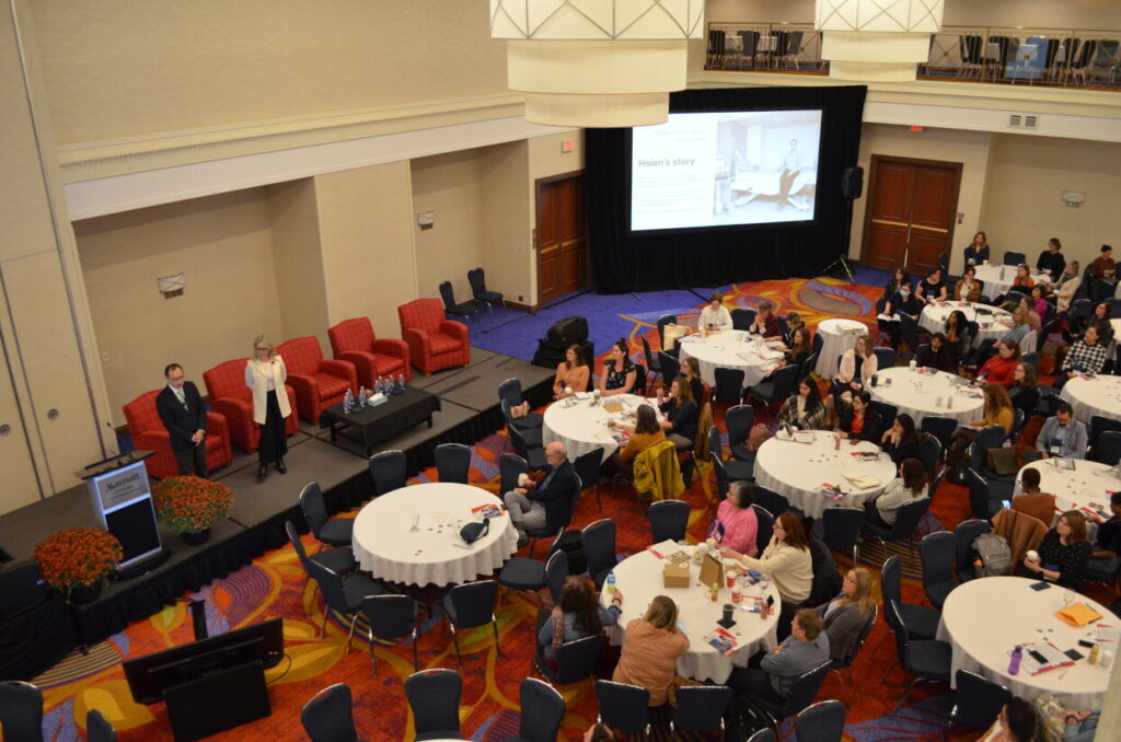 A photo from a hotel ballroom mezzanine with two people on a stage speaking to a large group of people seated around multiple circular tables.