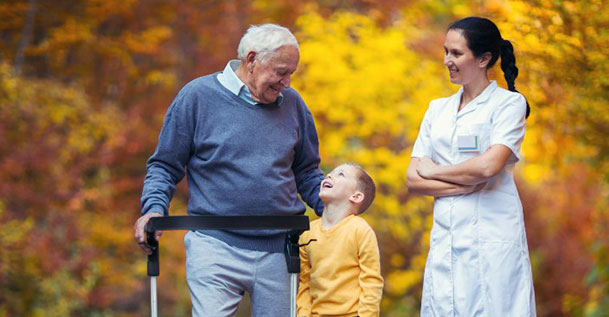 An elderly man with a walker walking with a young boy and a female nurse.