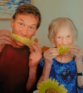 A man in a dark red t-shirt and an elderly woman in a blue patterned blouse enjoying corn on the cob together.